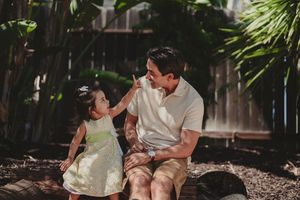 A father sits outdoors with his toddler daughter in a yellow dress, sharing a joyful moment among tropical greenery. Encinitas, CA