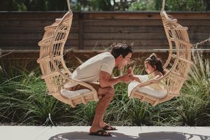 A father leans in to kiss his daughter sitting in a rattan crescent moon swing chair outdoors. Encinitas, CA