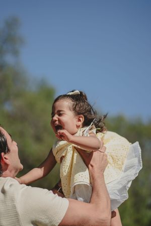 A joyful toddler girl in a yellow dress laughs as her father lifts her up against a clear blue sky. Encinitas, CA