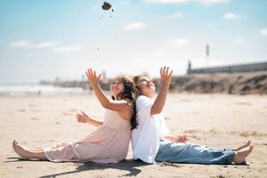 Siblings sitting back-to-back on a sunny beach, joyfully tossing sand into the air. Oceanside, CA
