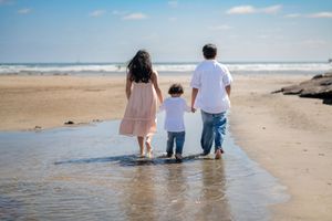 Family of three children walking hand-in-hand along a sunny beach shoreline, with ocean waves and blue sky in the background. Oceanside, CA