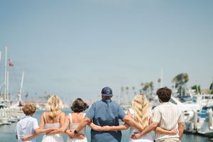 Family group photo from behind at a sunny marina, arms around each other, overlooking boats and blue sky. Oceanside, CA