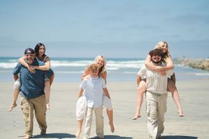 Happy family enjoying piggyback rides on a sunny beach, with blue skies and ocean waves in the background. Oceanside, CA