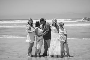 Black and white beach family portrait with five people embracing by ocean waves on a sunny day. Oceanside, CA