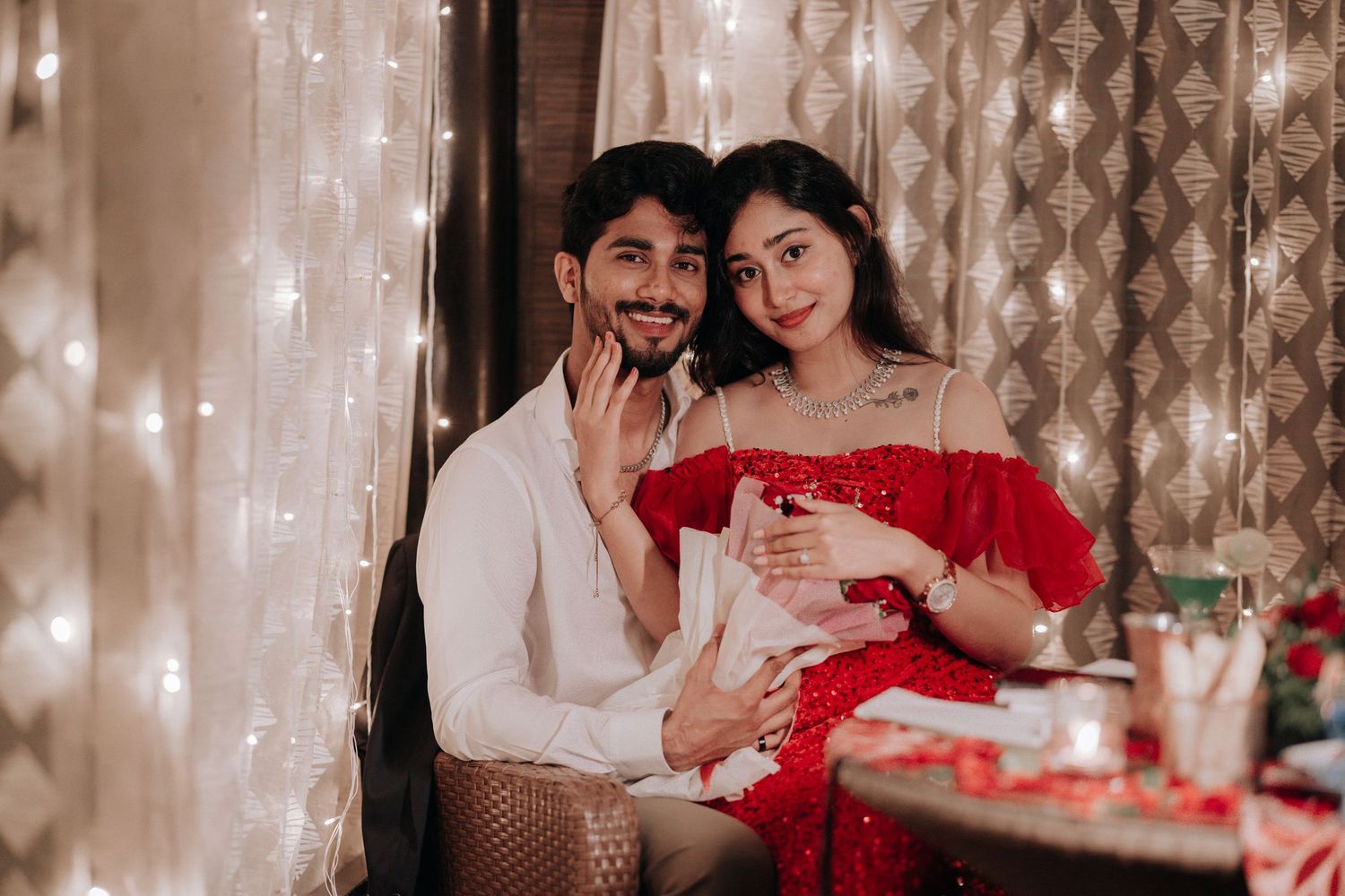 A couple in romantic attire poses together near fairy lights, the woman in red and man in white sharing a tender moment.