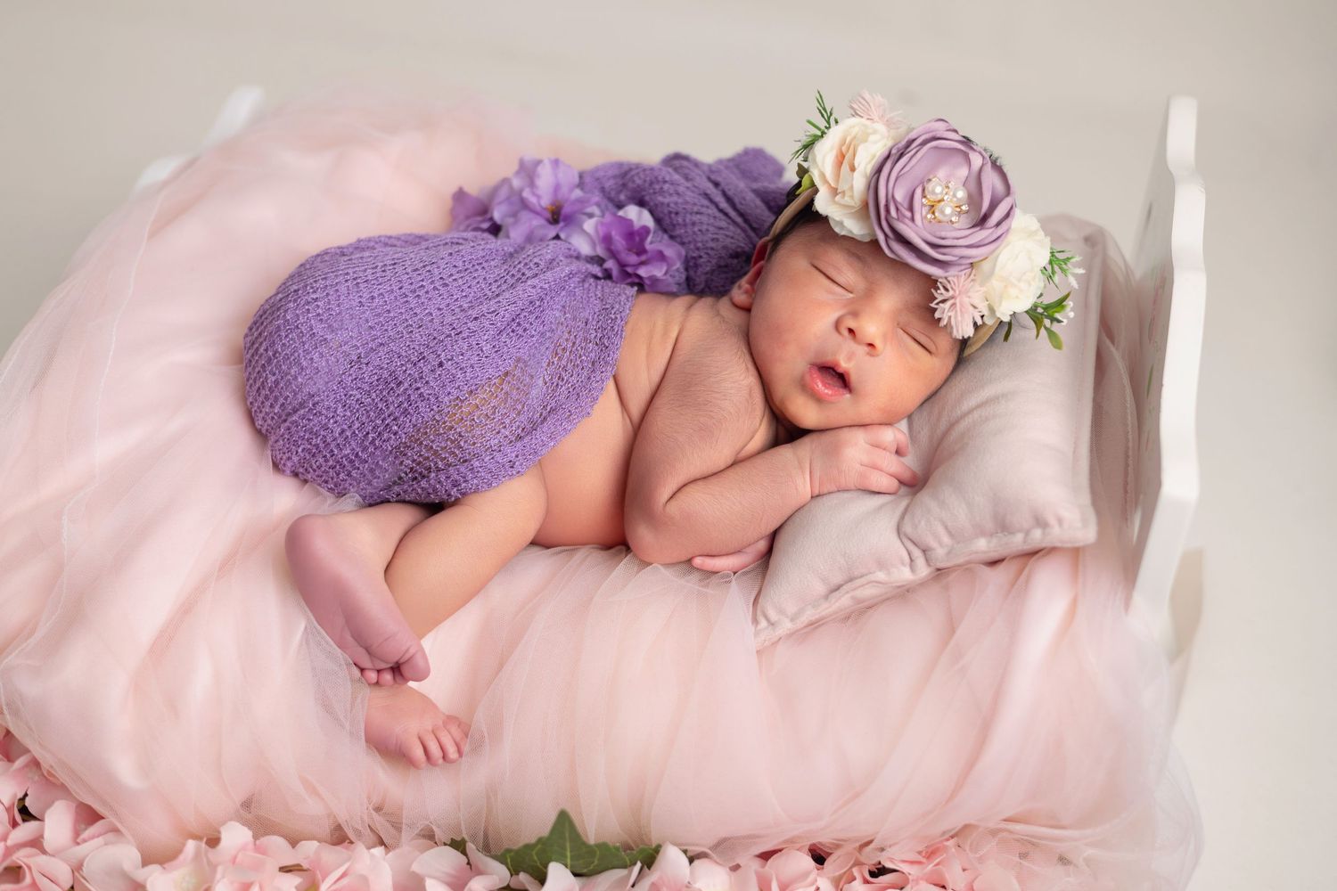 Sleeping newborn baby in purple knit outfit and floral headband resting on a tiny white bed with pink decor.