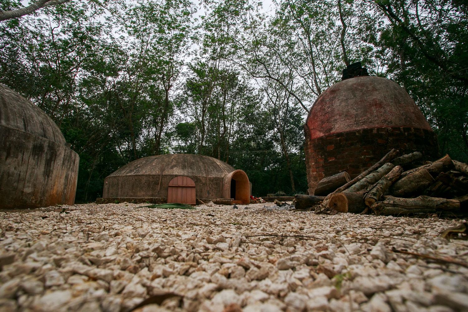 Traditional clay huts and domed structures stand against a backdrop of trees on rocky ground.
