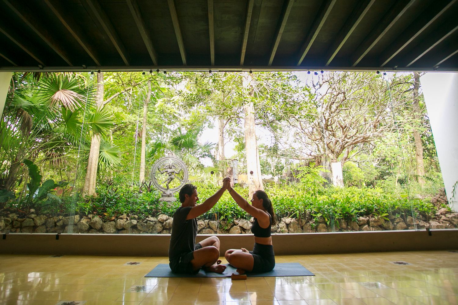 Two silhouettes sit together in a covered outdoor space overlooking tropical gardens.