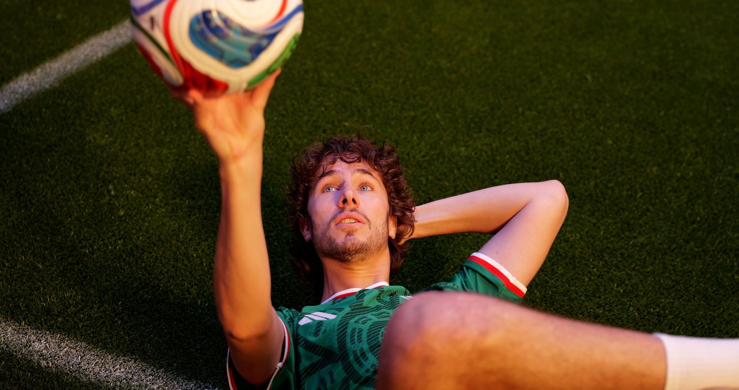 Soccer player in green jersey lying on field holding ball overhead under bright stadium lights.