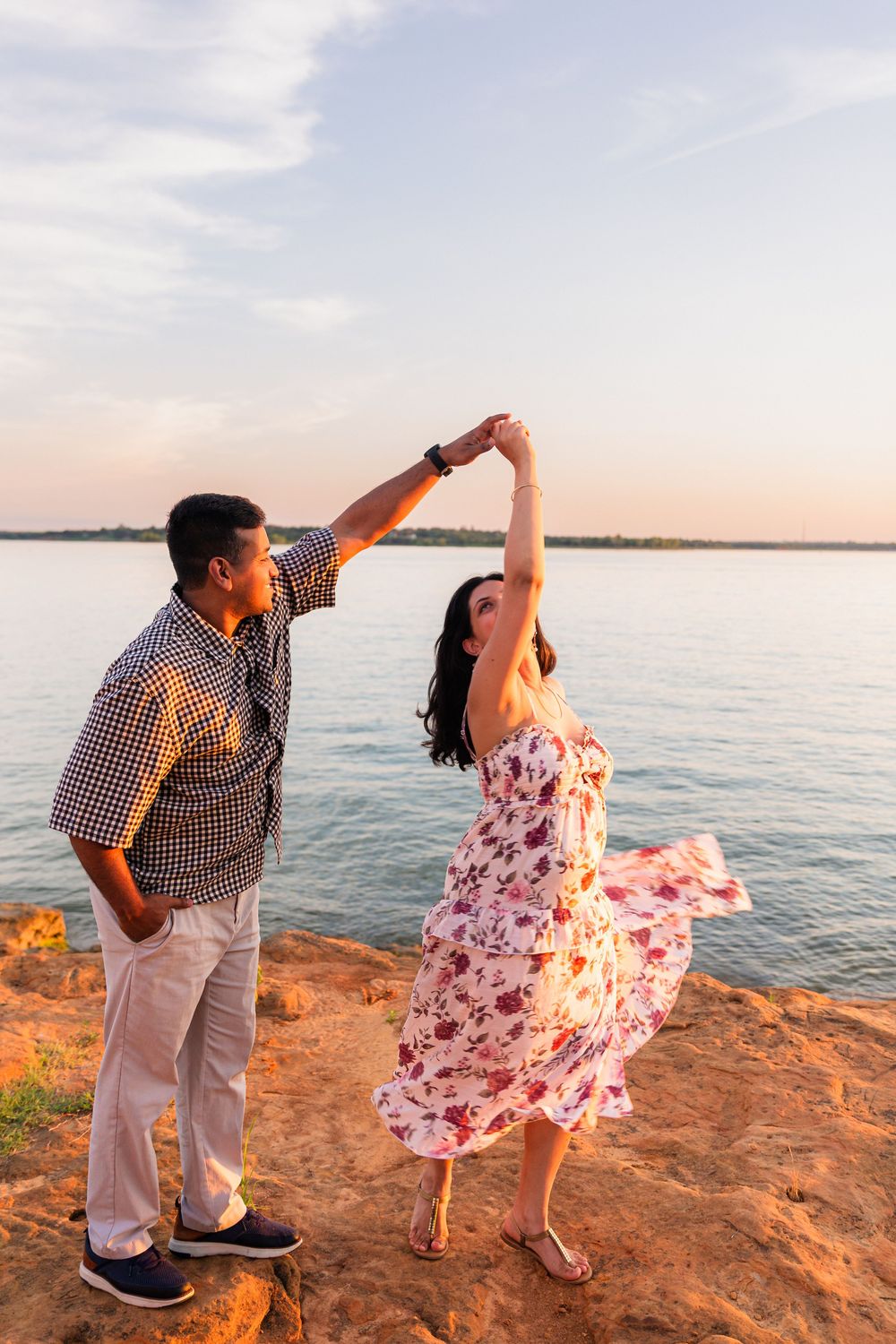 Couple dancing on rocky shore at sunset, woman in floral dress twirling, lake in background.