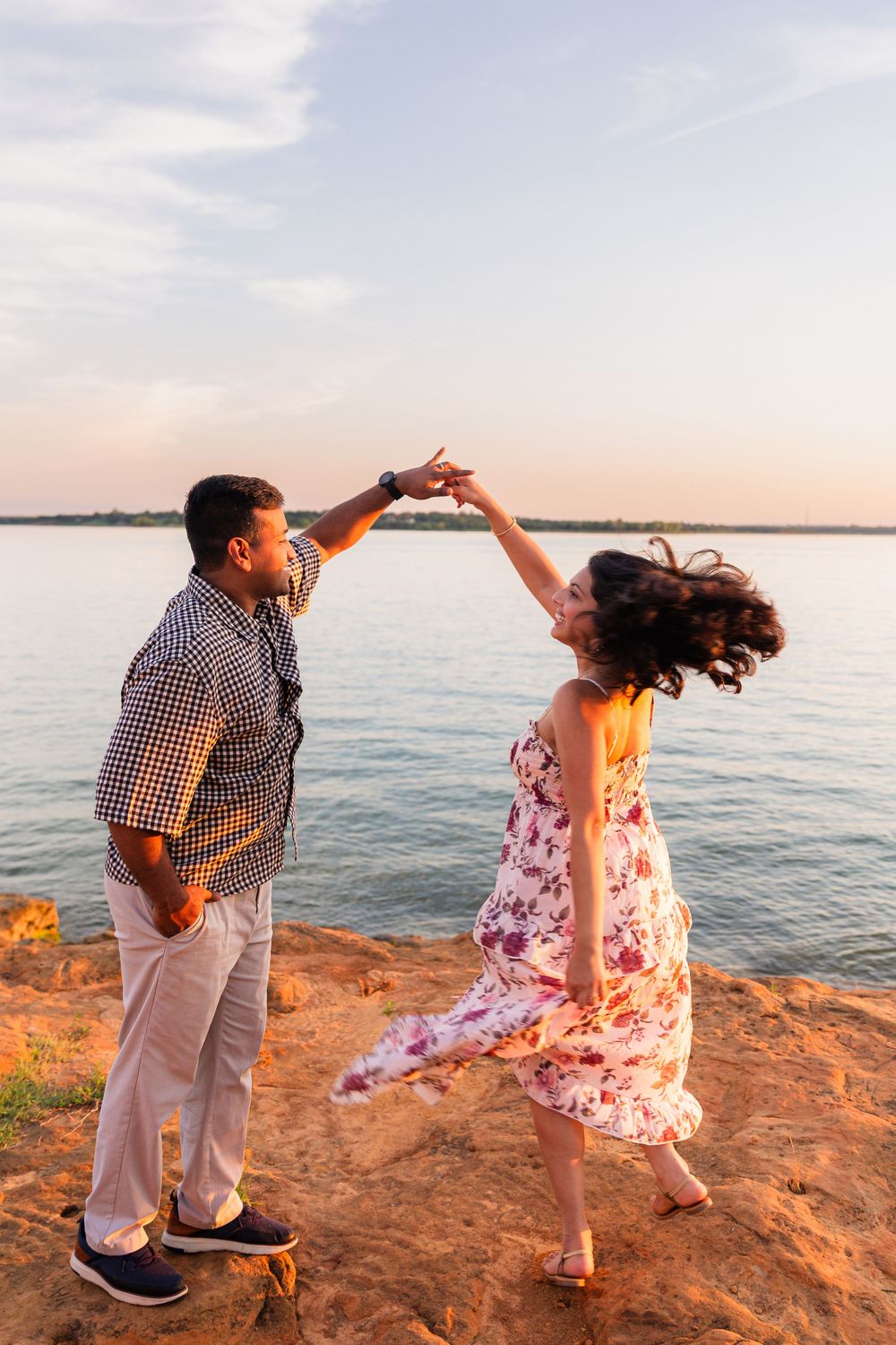 Couple dancing romantically on a rocky lakeside cliff at sunset, woman in floral dress being twirled by man in plaid shirt.