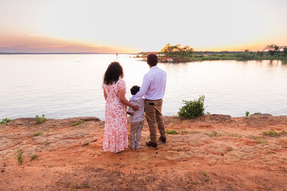 Family of three standing on red dirt cliff overlooking a serene lake at golden sunset.