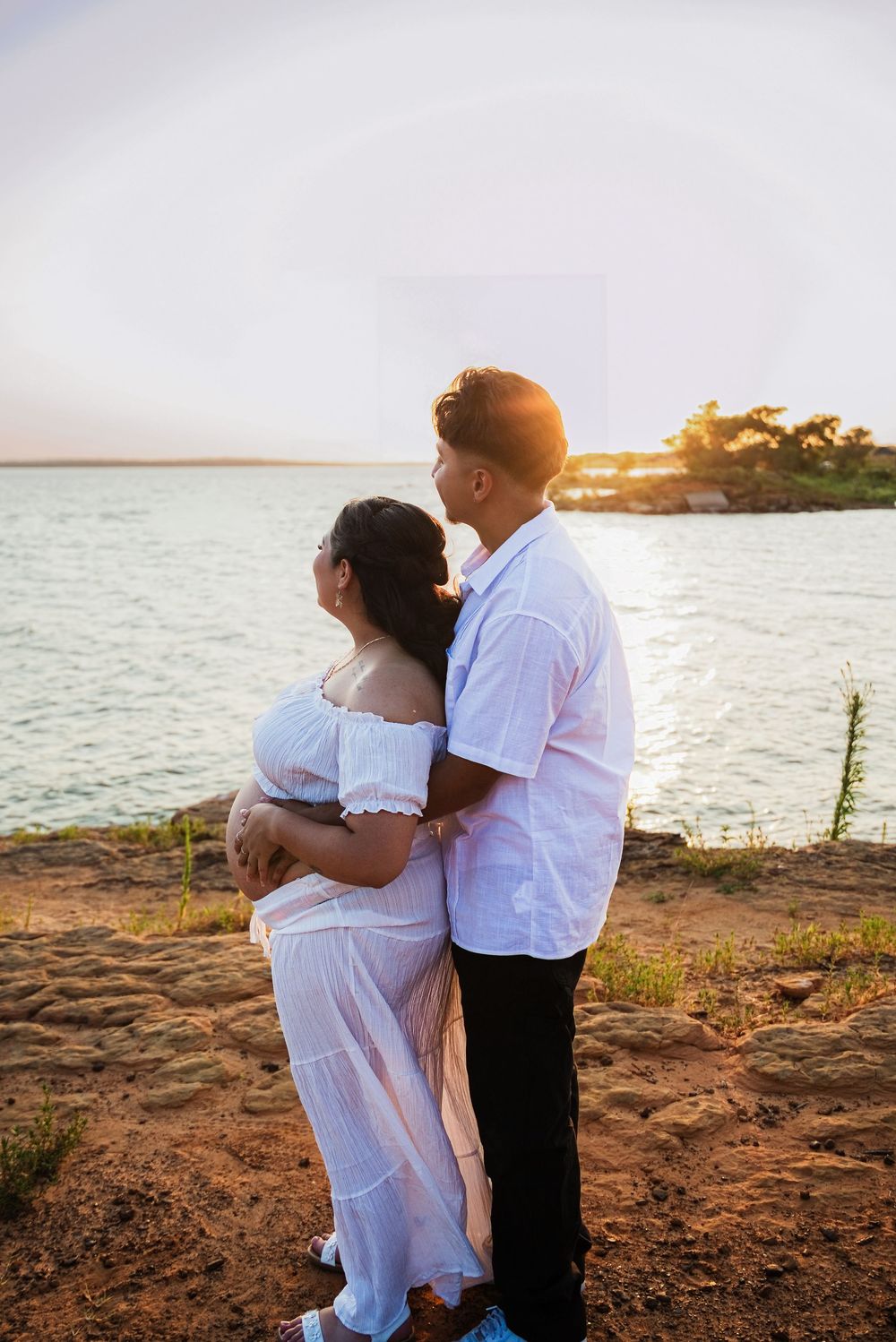 Couple embracing at golden hour by the water, woman in white dress, man in white shirt, standing on rocky shore.