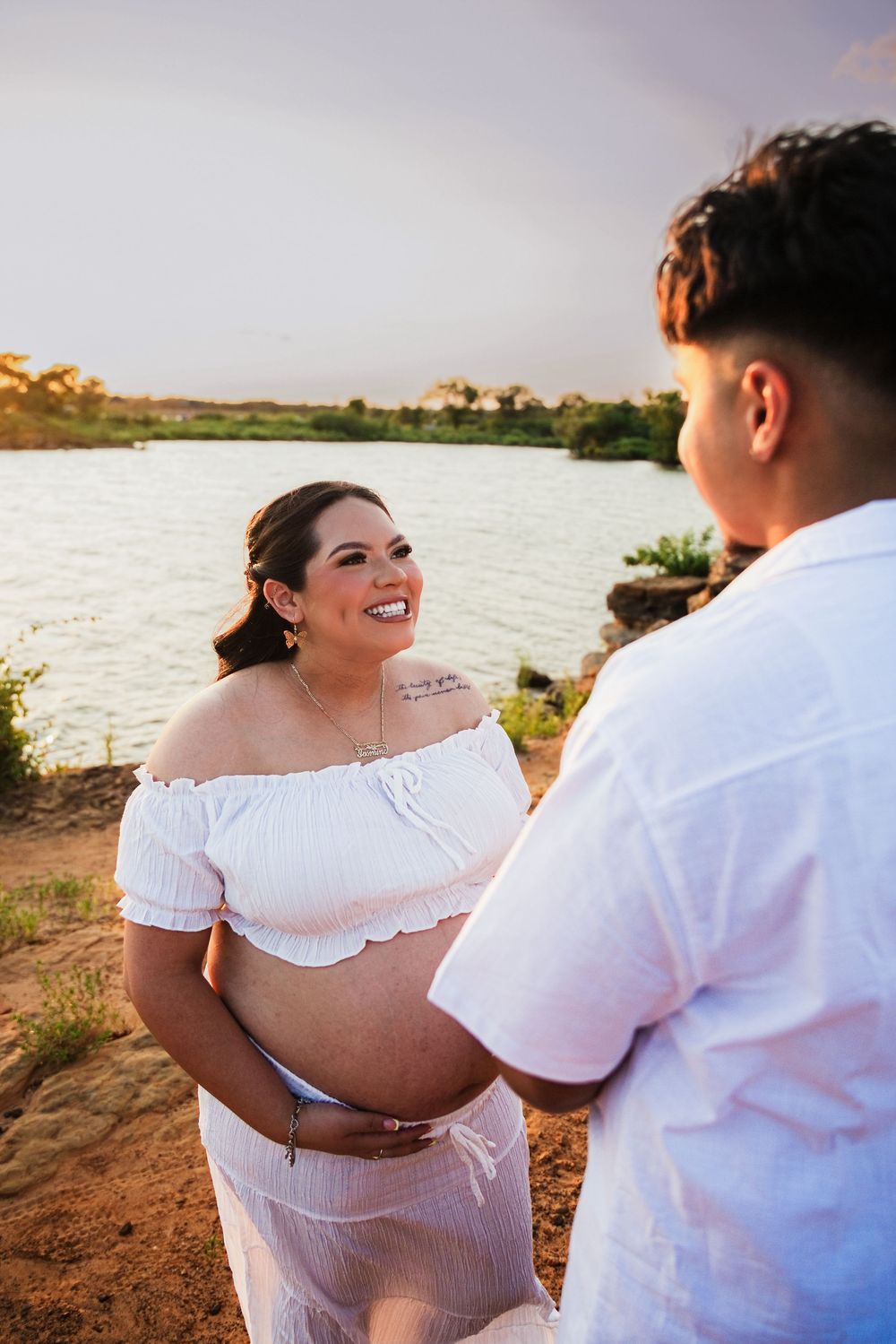 Pregnant woman in white off-shoulder dress smiles at partner outdoors near a lake during golden hour maternity photoshoot.