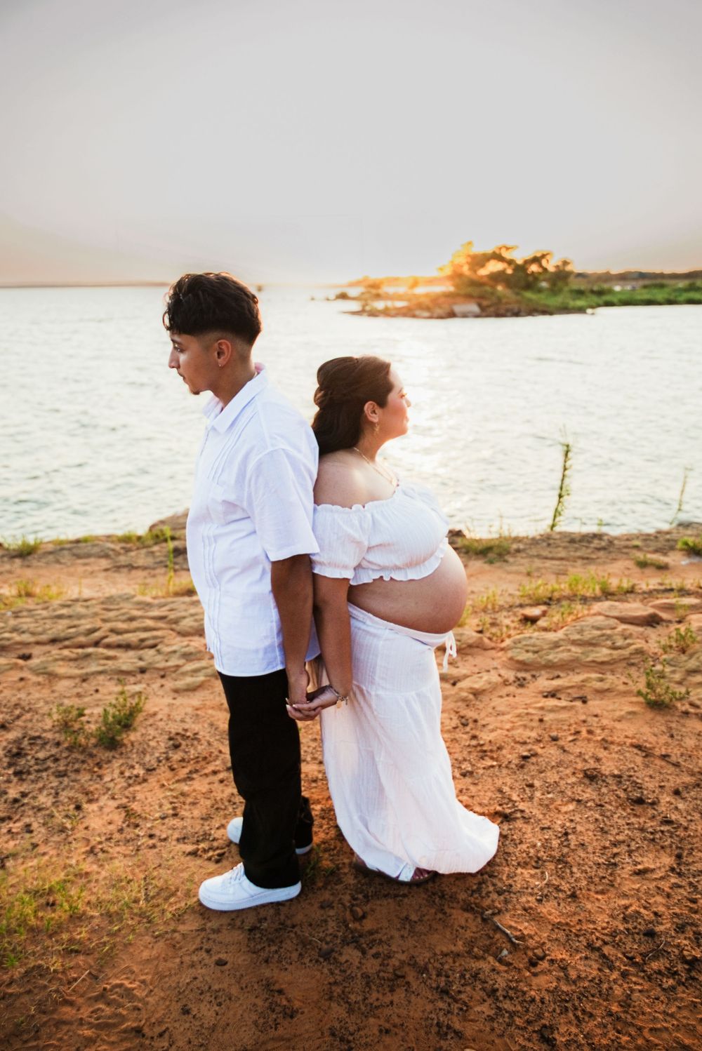 Pregnant couple in white outfits holding hands back-to-back on red earth by a serene lake at golden hour.