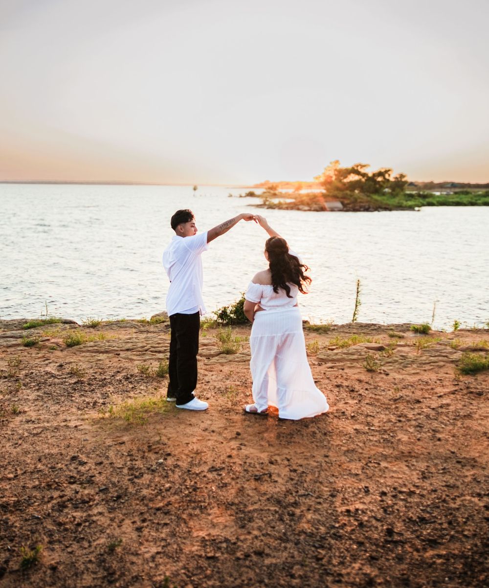 Couple in white dancing by the water at sunset on rocky shore.