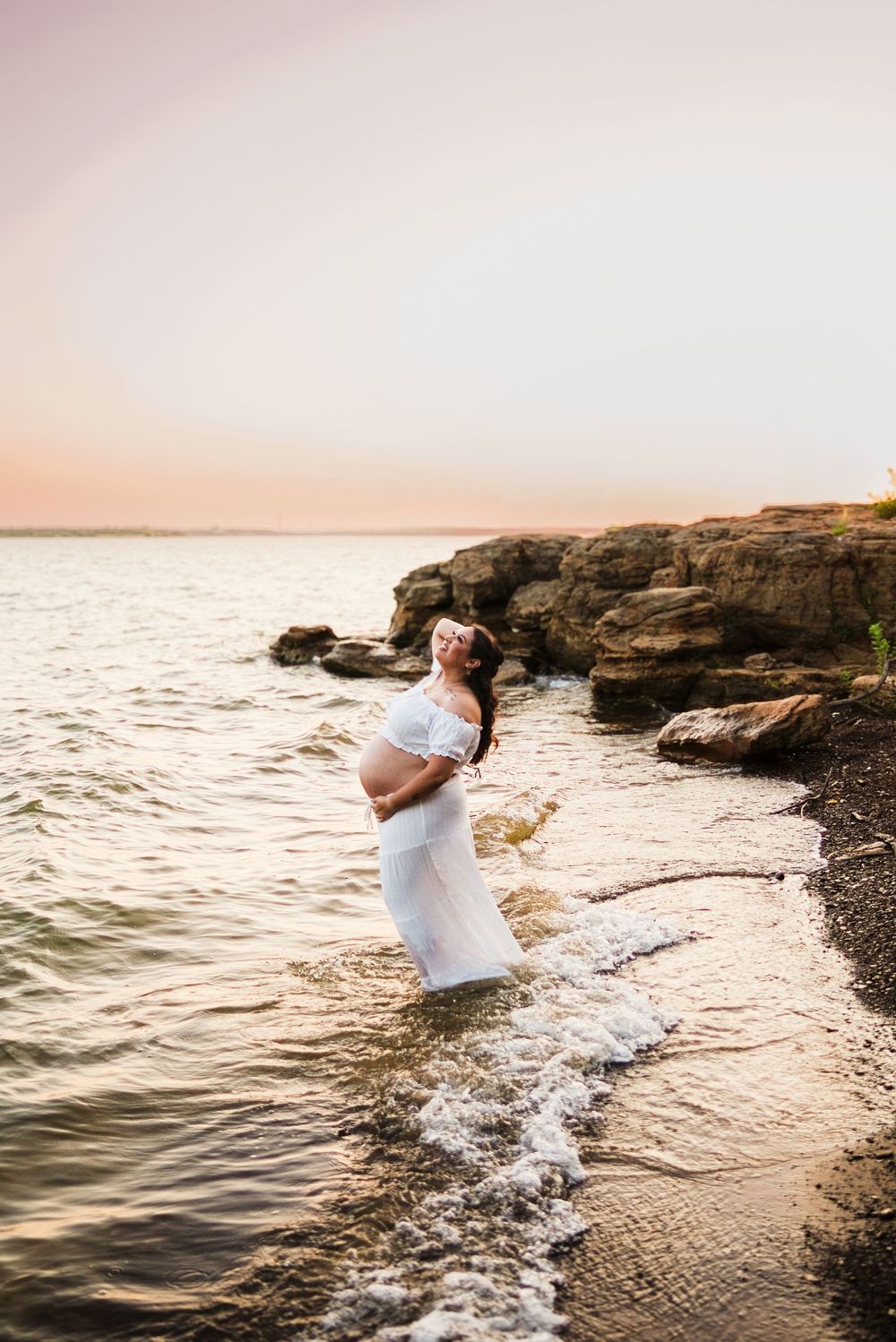 Pregnant woman in white dress standing in ocean waves at sunset near rocky shore.