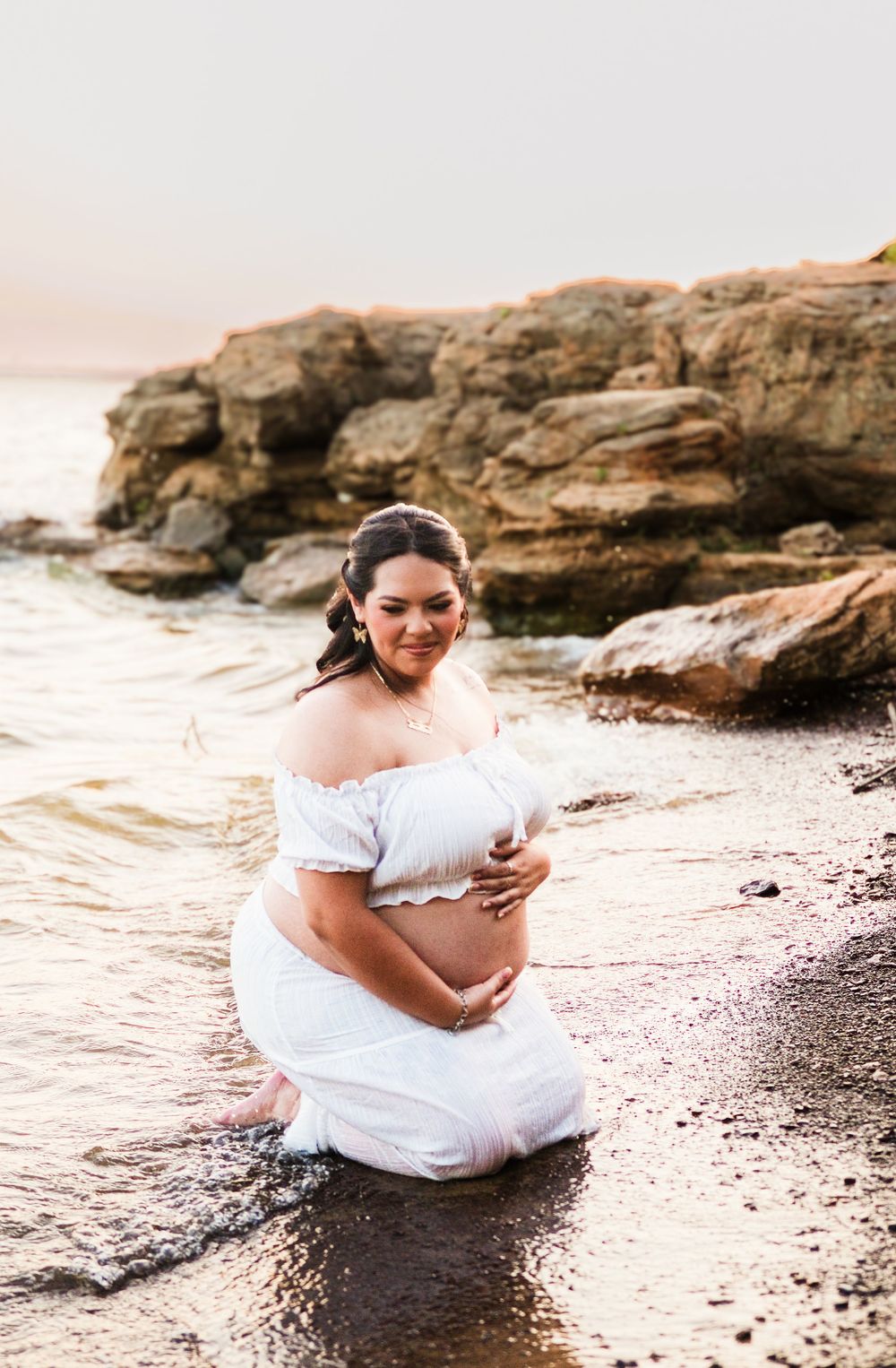 Pregnant woman in white off-shoulder dress kneeling in shallow ocean water near rocks during golden hour maternity photo session.