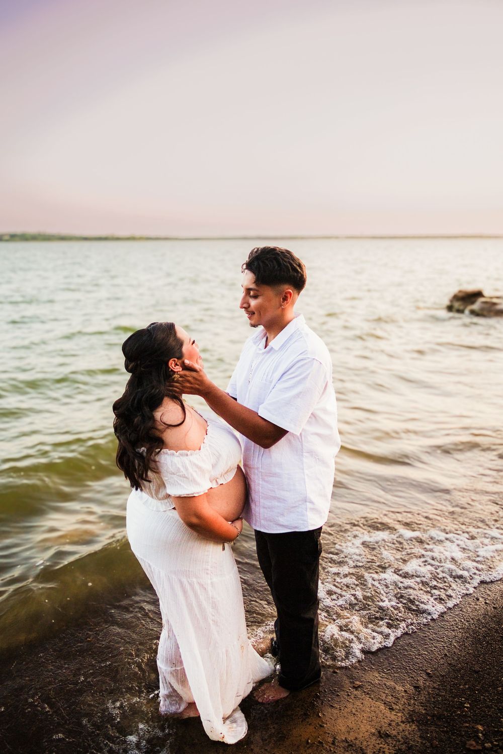 Couple sharing a tender moment at the beach during sunset, woman in white maternity dress, man in white shirt.