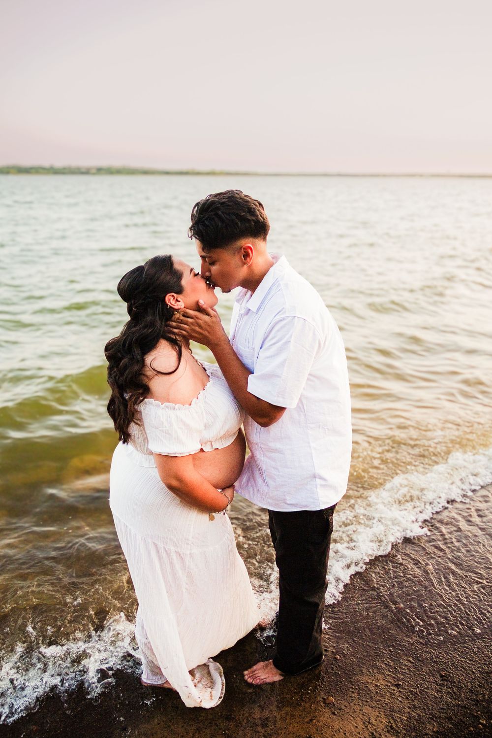 Pregnant couple sharing a romantic kiss while standing in ocean waves, woman in white maternity dress, beach maternity photo.
