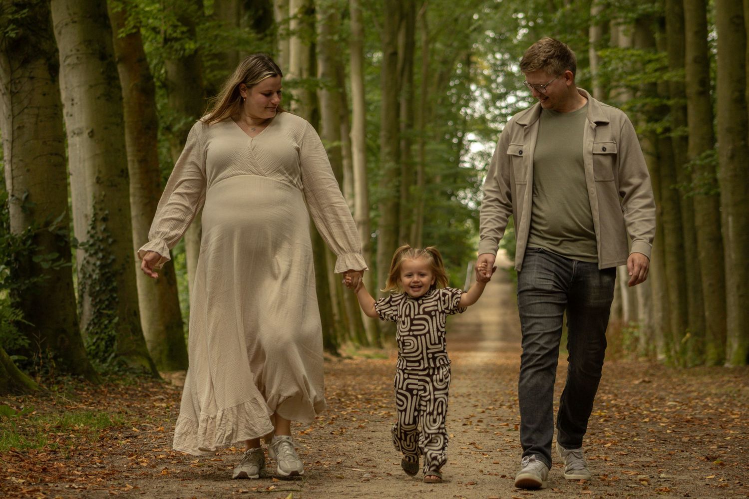 Twee ouders lopen hand in hand met hun lachende kind over een bospad.