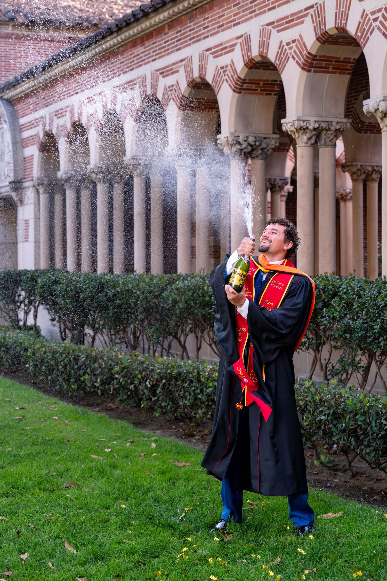 Graduate in black robe celebrating with champagne spray outside ornate university building with arched colonnade.
