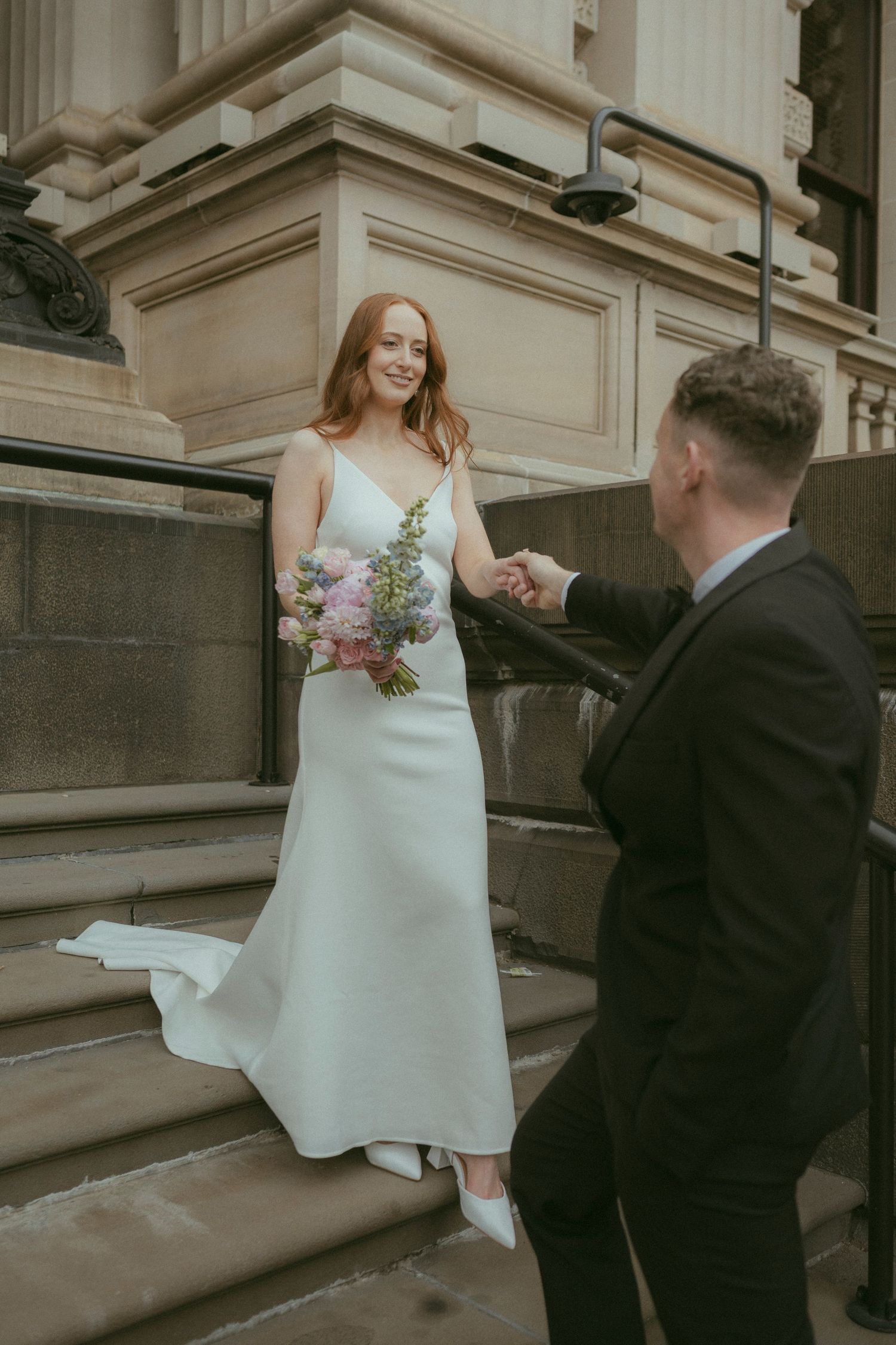 Bride in white gown holds bouquet on stone steps while groom in black suit holds her hand at outdoor wedding.