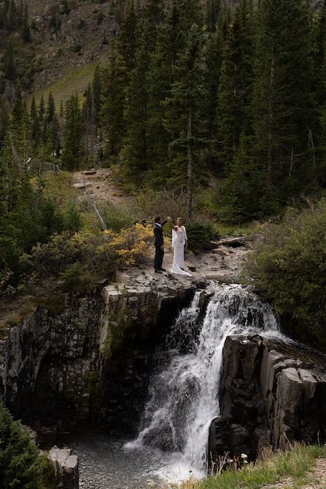 Bride and groom stand atop a rocky waterfall surrounded by pine trees in a stunning mountain elopement ceremony.
