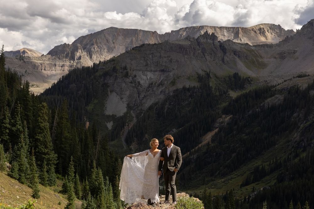 Bride and groom stand on rocky mountain overlook with dramatic peaks and pine forests in Colorado elopement wedding.