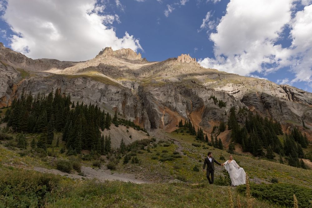 Couple in wedding attire walking through alpine meadow with dramatic rocky mountain peaks and evergreen forest backdrop.