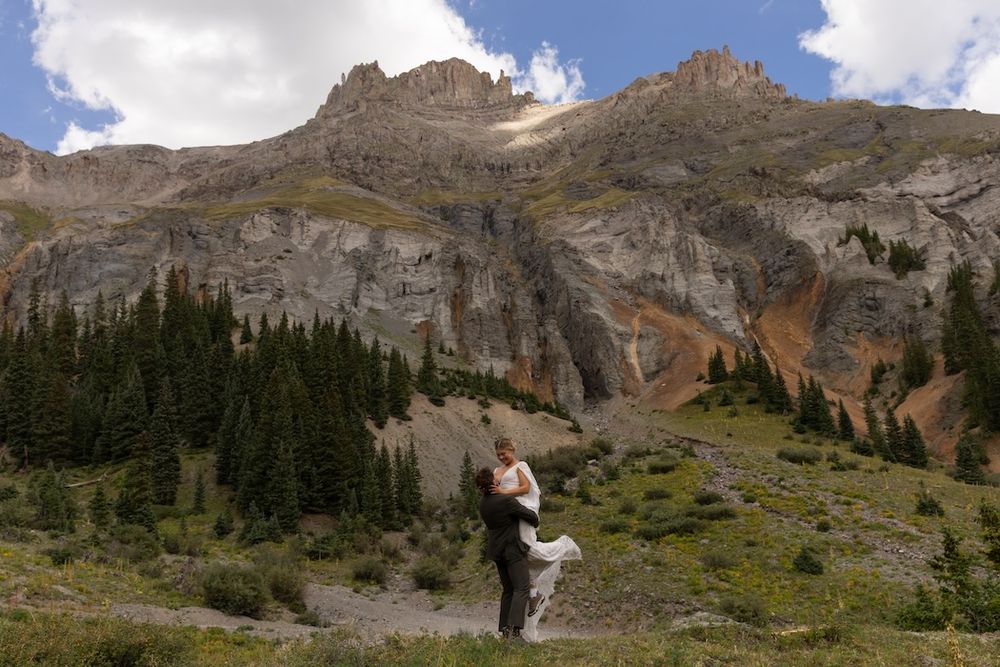 Couple embracing in wedding attire with stunning Rocky Mountain backdrop and evergreen trees under a partly cloudy sky.