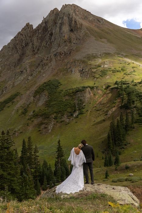 Bride and groom stand on a rock overlooking a stunning Colorado mountain landscape with green slopes and evergreen trees.
