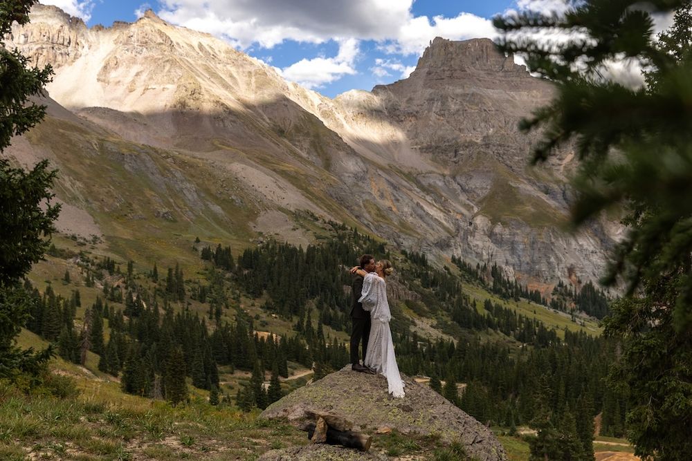Couple embracing on a rocky ledge with stunning mountain peaks and evergreen forest backdrop on a sunny day.