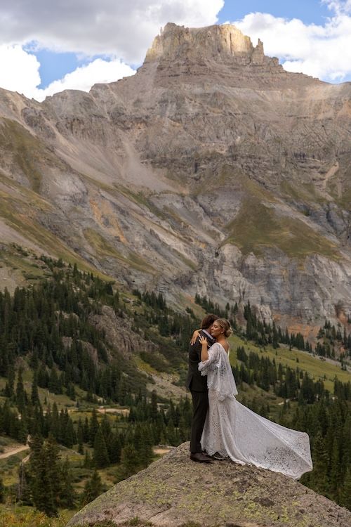Bride and groom embrace on a rocky ledge with stunning mountain peaks and pine forest backdrop.