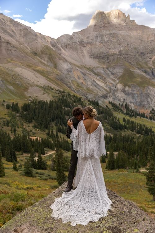Bride in lace gown and groom stand on rocky overlook with mountain peaks and pine forest backdrop.
