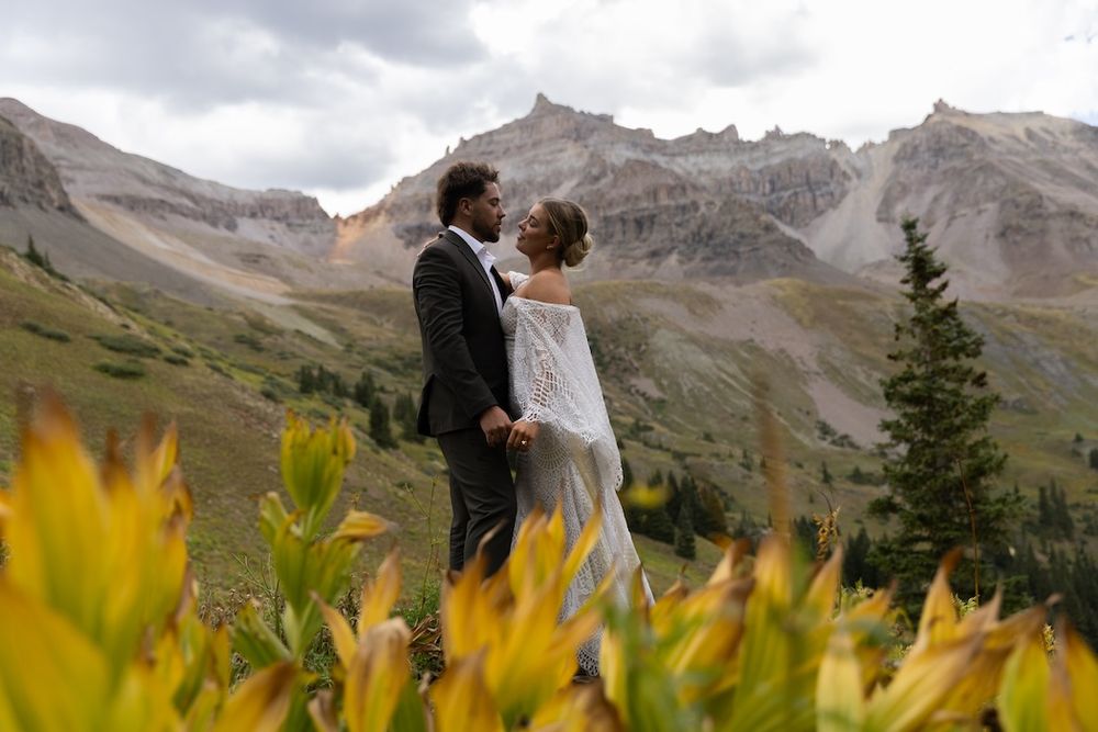 Couple in wedding attire embracing in a mountain meadow with yellow wildflowers and rocky peaks in the background.