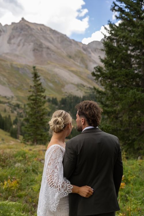 Bride and groom stand together gazing at a stunning Rocky Mountain landscape surrounded by evergreen trees.
