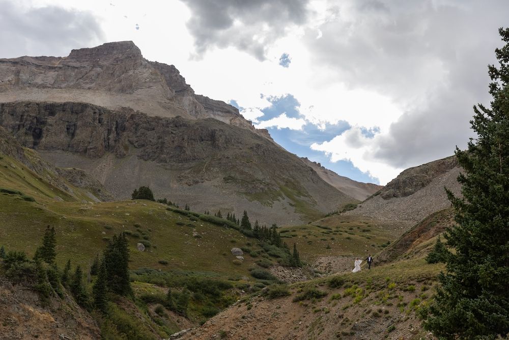 Scenic alpine valley with lush green slopes, scattered evergreen trees, and towering rocky mountain peaks under cloudy sky.
