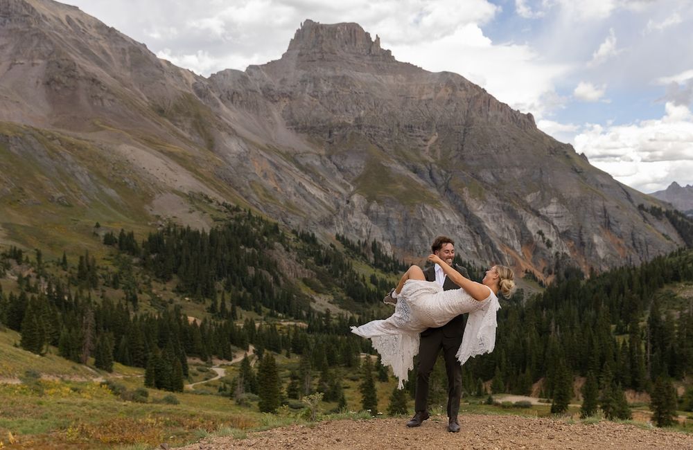 Groom lifting bride in arms with stunning Rocky Mountain peaks and pine trees backdrop.