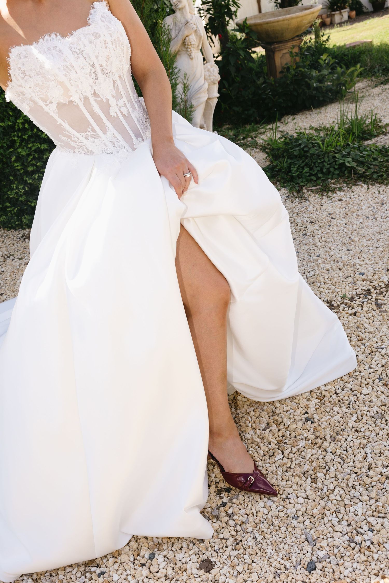 Bride in white lace corset wedding gown with thigh slit, lifting skirt to reveal burgundy pointed flats on gravel path.