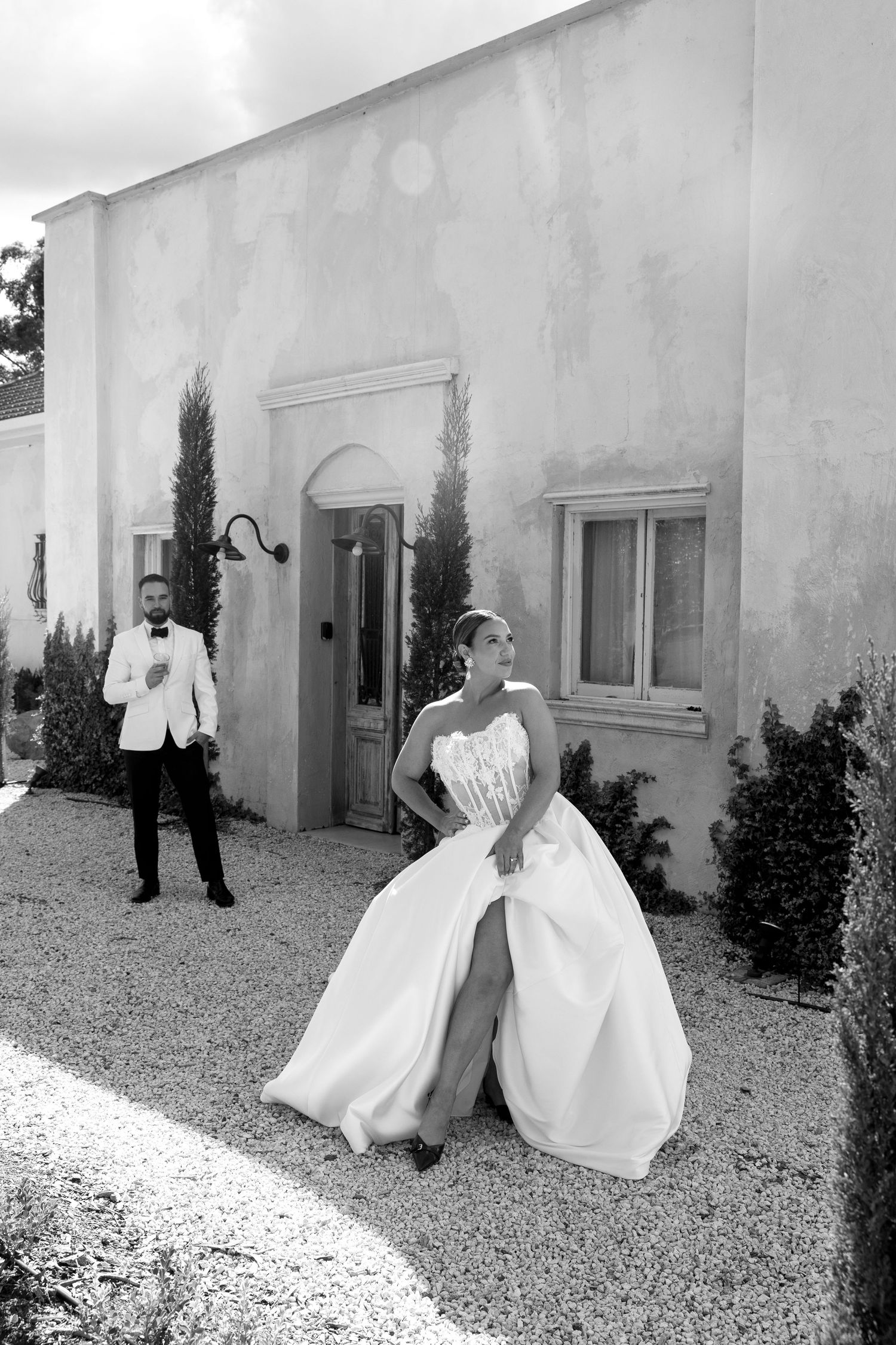 Black and white photo of a bride in a strapless gown and groom in white tuxedo outside a rustic villa.