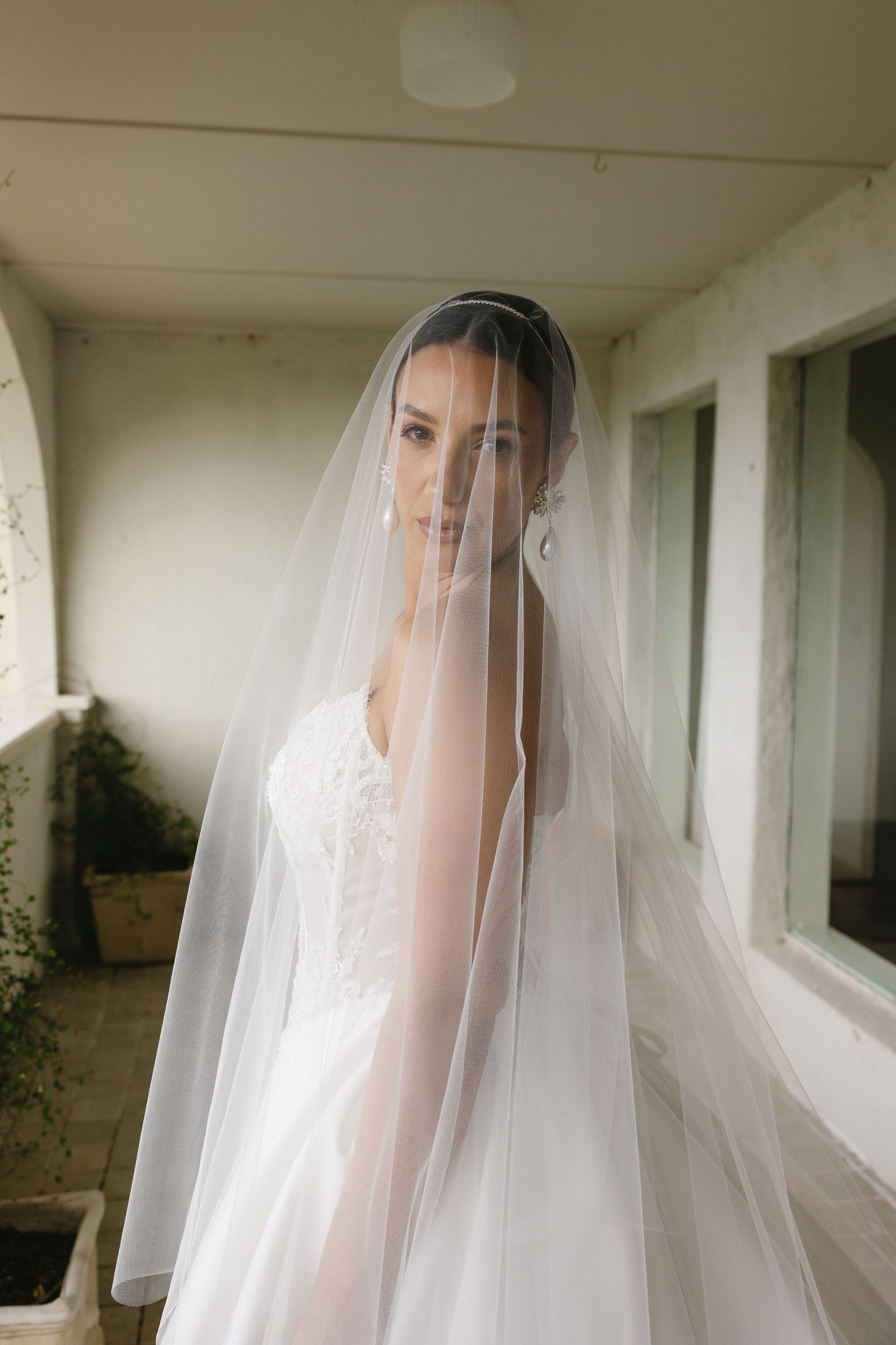 Elegant bride wearing a flowing white veil over her face and lace wedding dress, standing in a softly lit indoor space.
