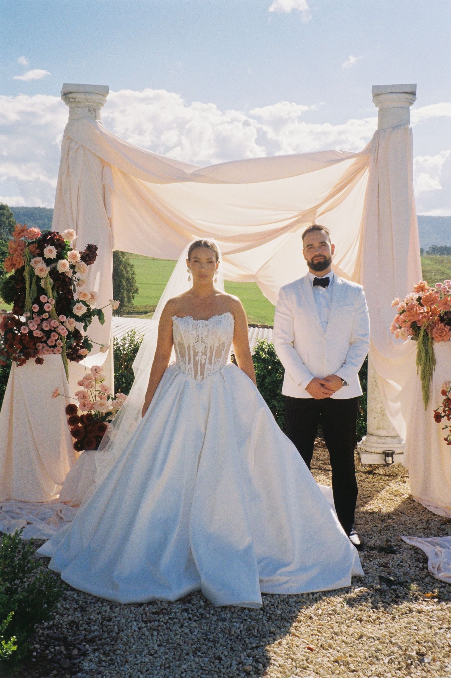 Bride in strapless ballgown and groom in white tuxedo stand at outdoor wedding altar with floral arrangements.