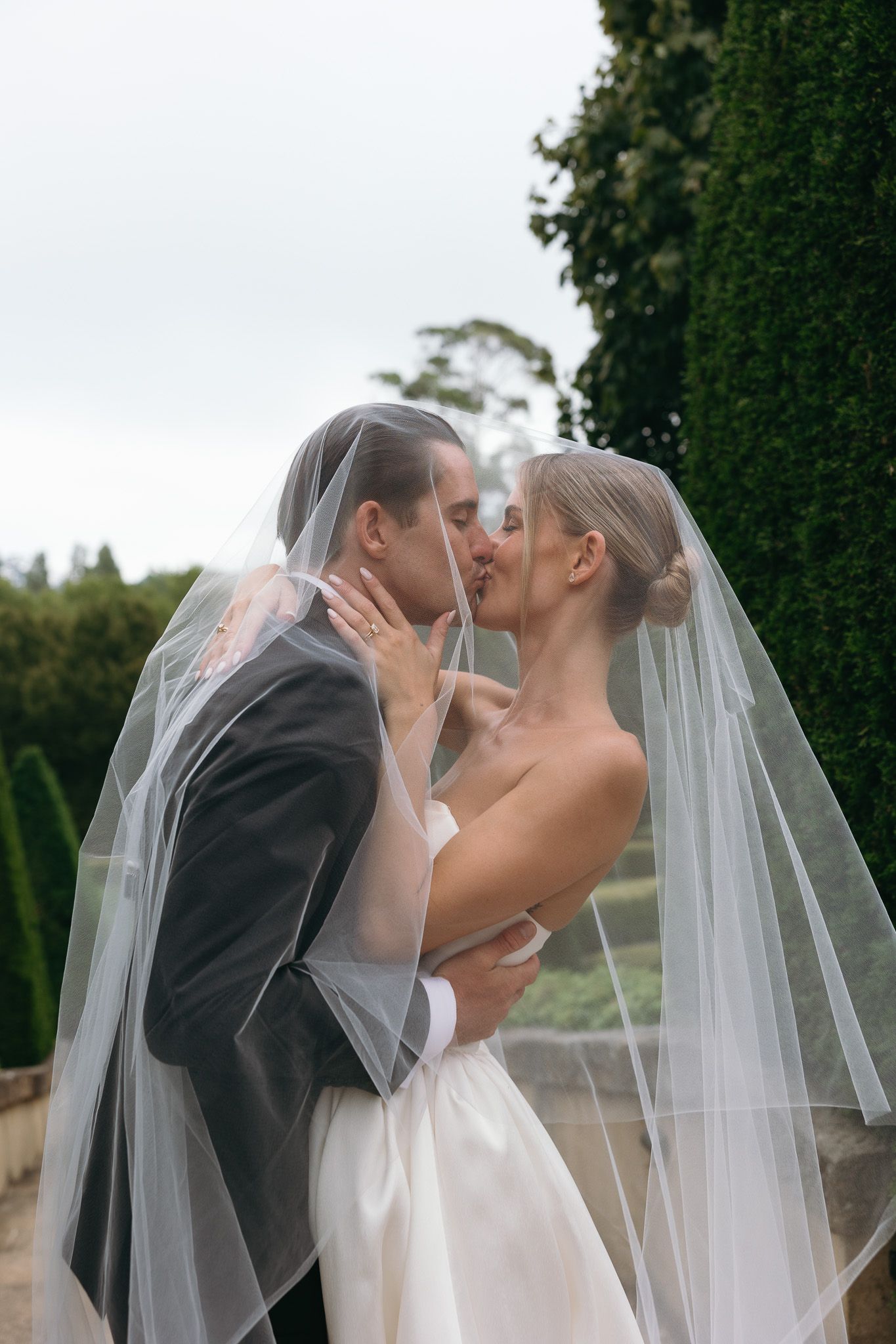 Bride and groom share a romantic kiss under a flowing wedding veil outdoors, surrounded by lush greenery.