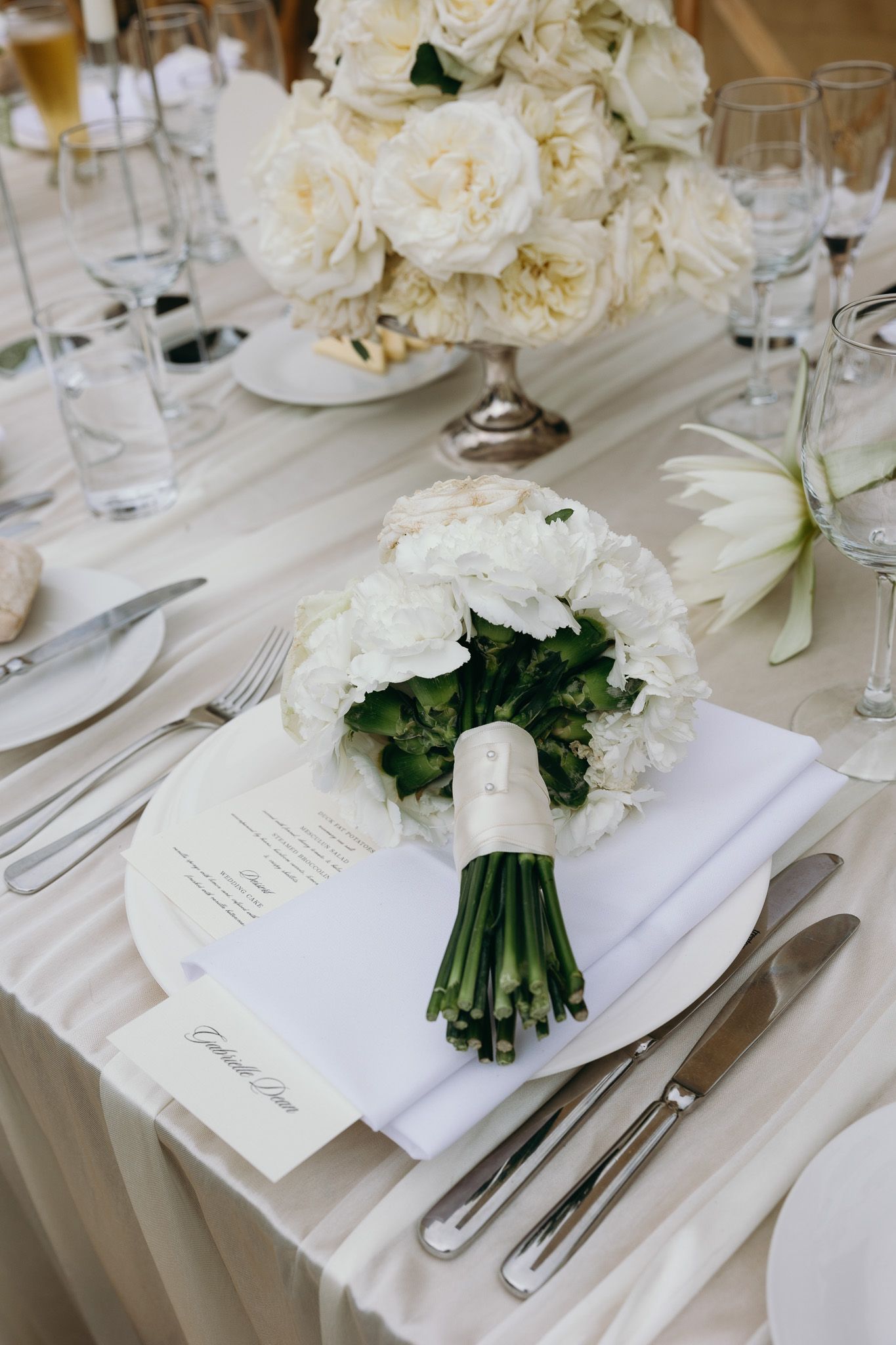 Elegant wedding table setting with white floral bouquet, menu card, silverware, and ivory rose centerpiece on white linen.