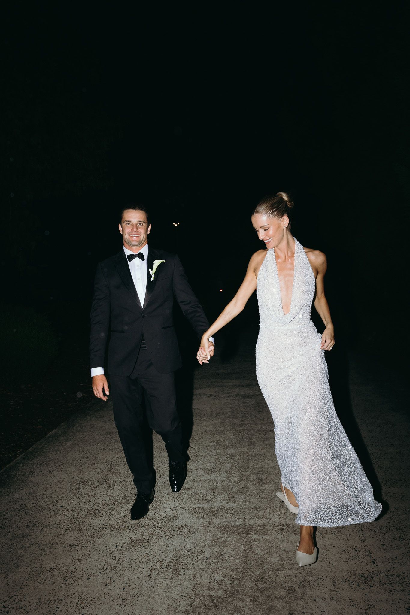 Bride and groom walking hand in hand at night, she in a white halter gown, he in a black tuxedo.