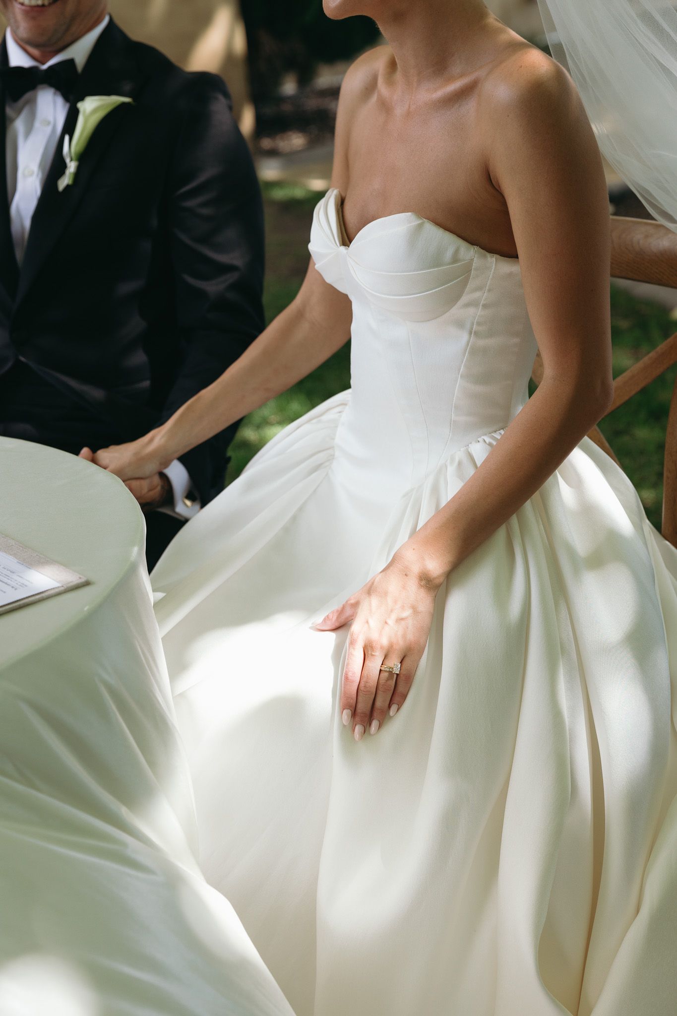 Bride in strapless white ball gown wedding dress with sweetheart neckline, standing beside groom in black tuxedo outdoors.