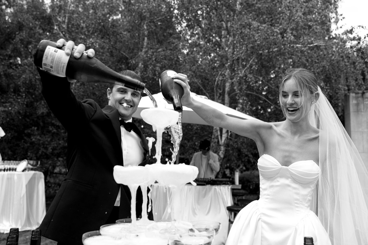 Black and white photo of a bride and groom joyfully pouring champagne over a tower of glasses at their wedding reception.