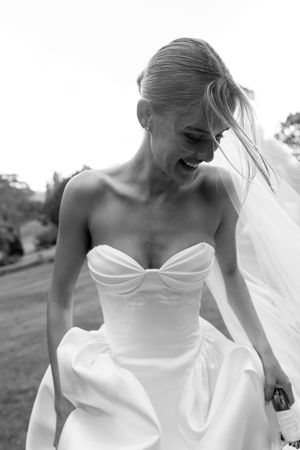 Laughing bride in strapless white wedding gown with veil blowing in wind, black and white outdoor photo.