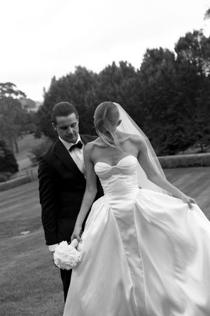 Black and white photo of a bride and groom walking hand in hand outdoors, bride in strapless gown with veil, groom in tuxedo.